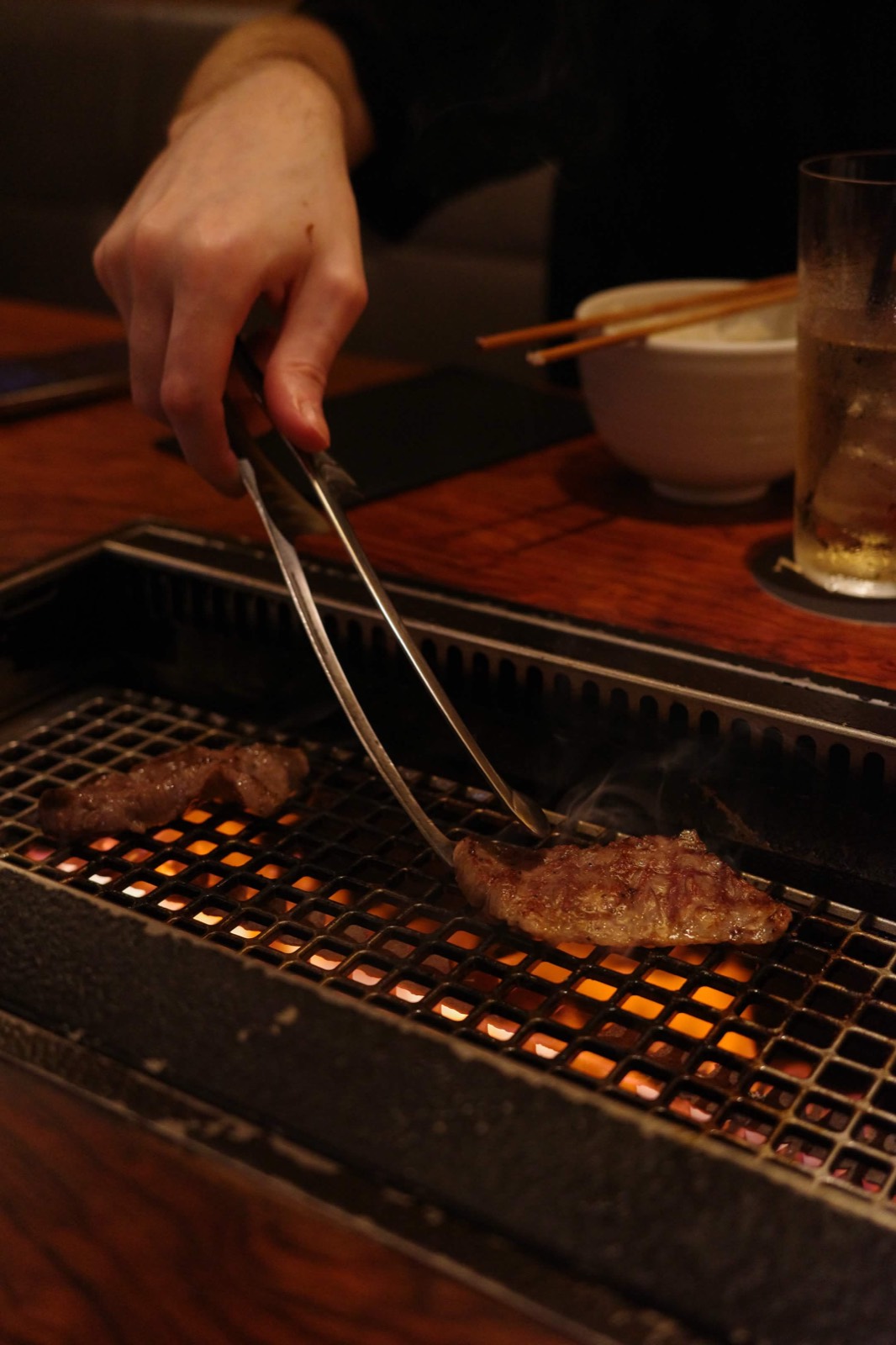 Hand with tongs turning beef over a glowing charcoal grill set into a wooden table.