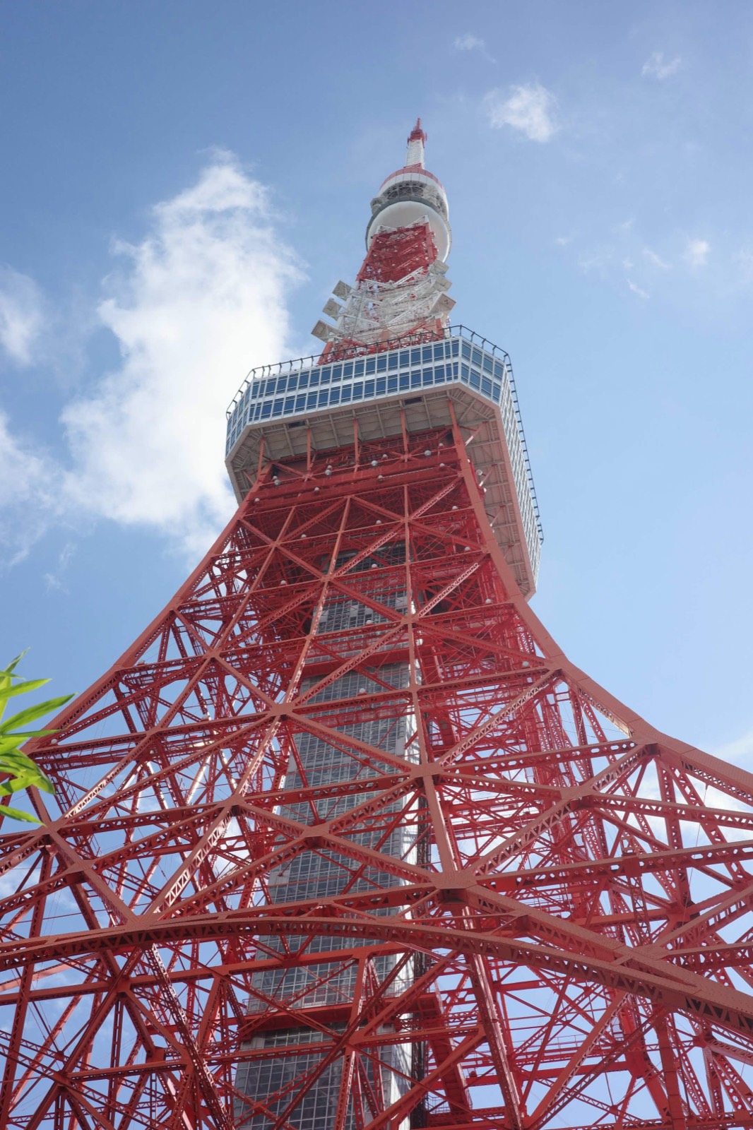 Tokyo Tower's red lattice steelwork seen from directly below against a blue sky with drifting clouds.