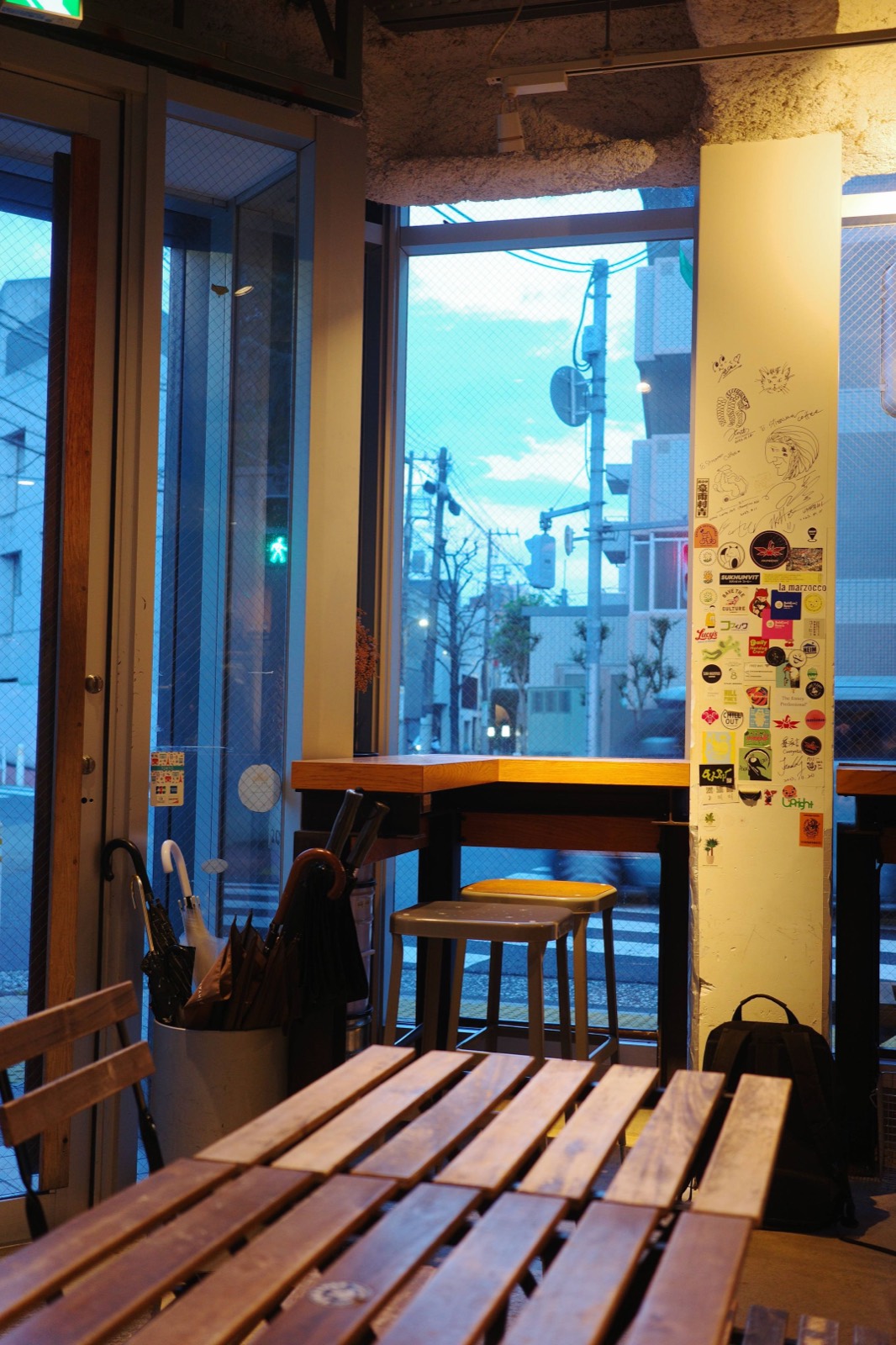 Corner of a café at dusk, a sticker-covered pillar beside window counters looking out on a quiet street.